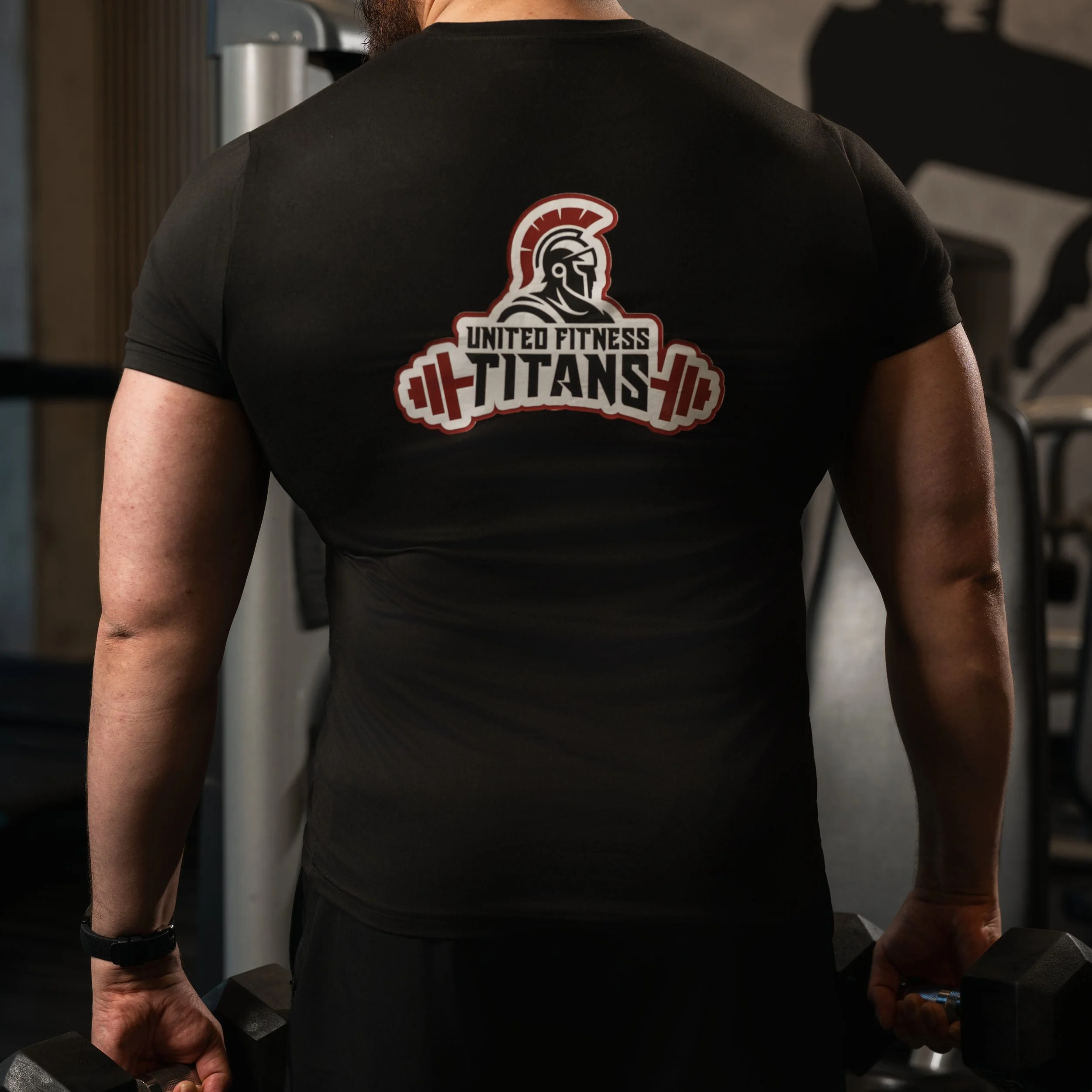 Person in a black T-shirt with "United Fitness Titans" logo, holding dumbbells in a gym.