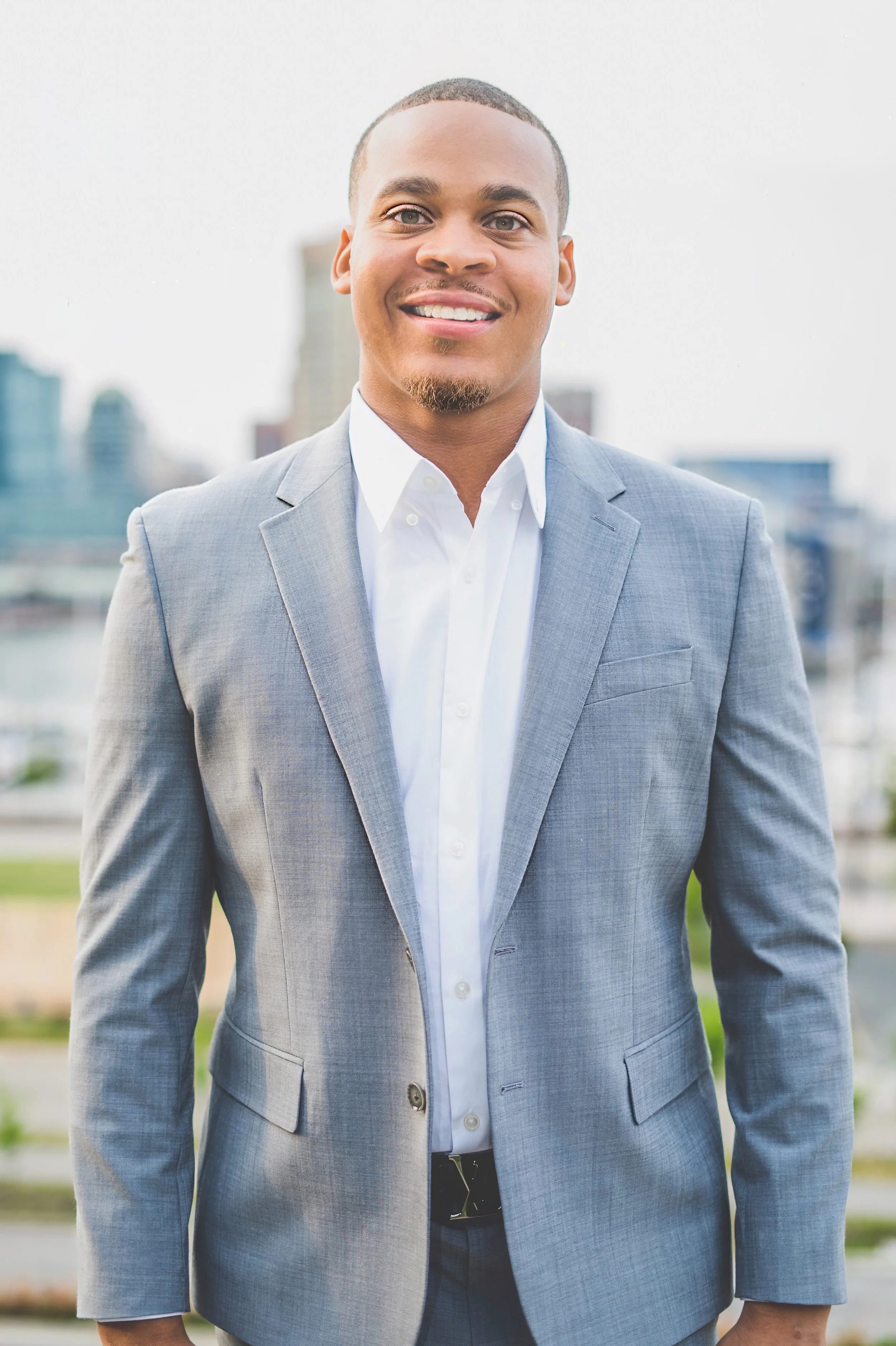 A man wearing a light gray suit and white shirt, smiling outdoors with a cityscape in the background.
