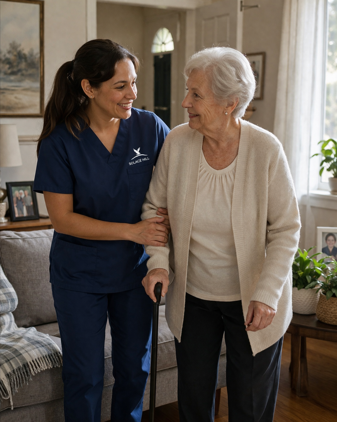 A caregiver in navy blue scrubs helping an elderly woman walk with a cane in a living room with sunlight and houseplants.