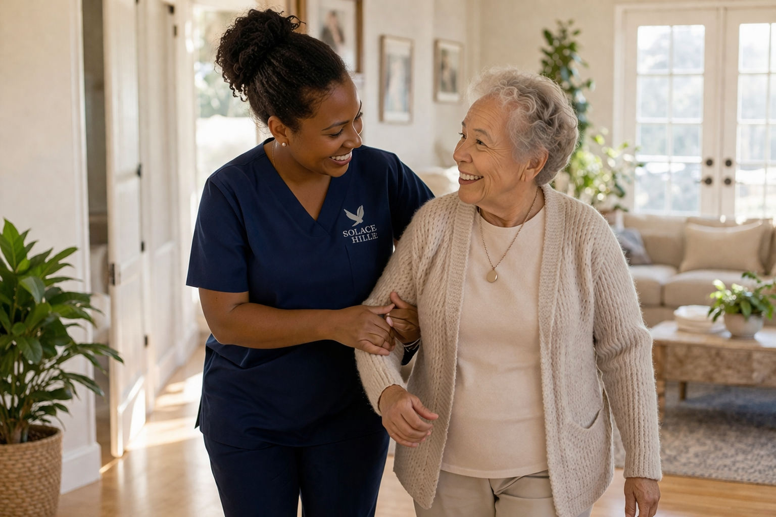 A young woman healthcare worker in navy scrubs smiling and holding hands with an elderly woman in a cozy living room.