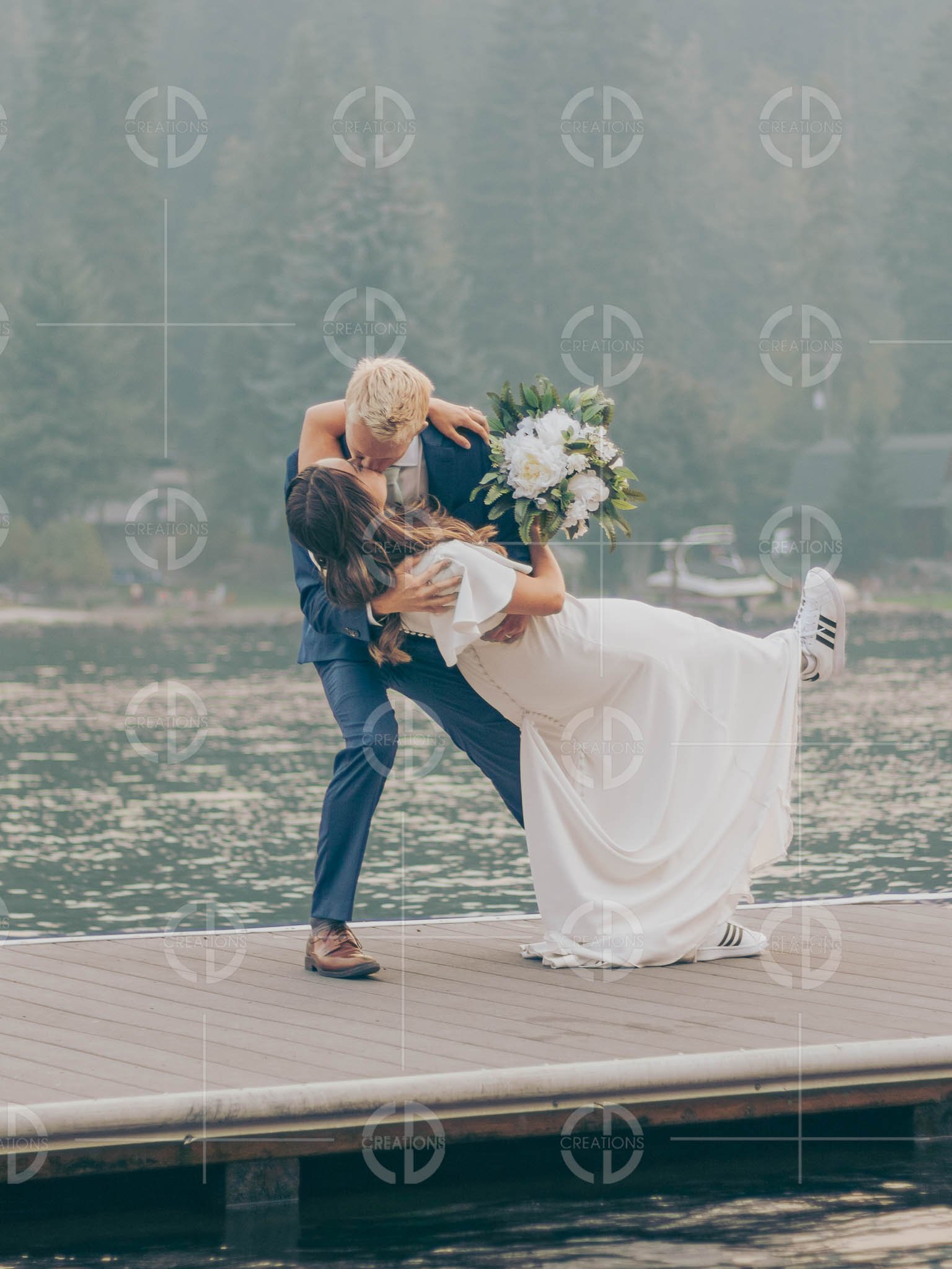 A newlywed couple on a wooden dock by a lake, with the groom dipping the bride while she holds a bouquet of white flowers, surrounded by a calm lake and forested background.