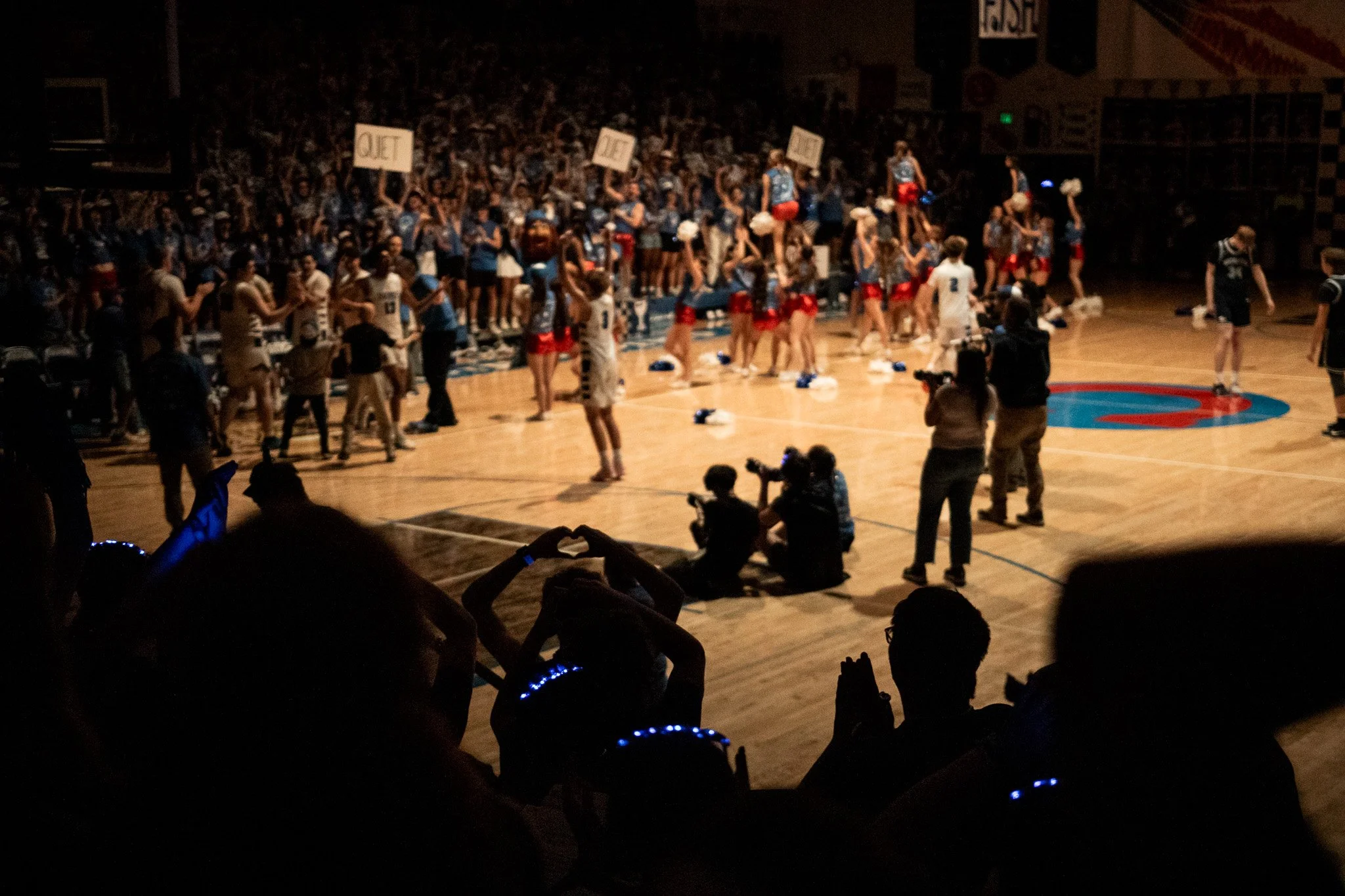 A large indoor basketball arena filled with cheerleaders in blue jerseys and red skirts, holding signs, and spectators, some filming or taking photos, during a cheerleading event or competition.