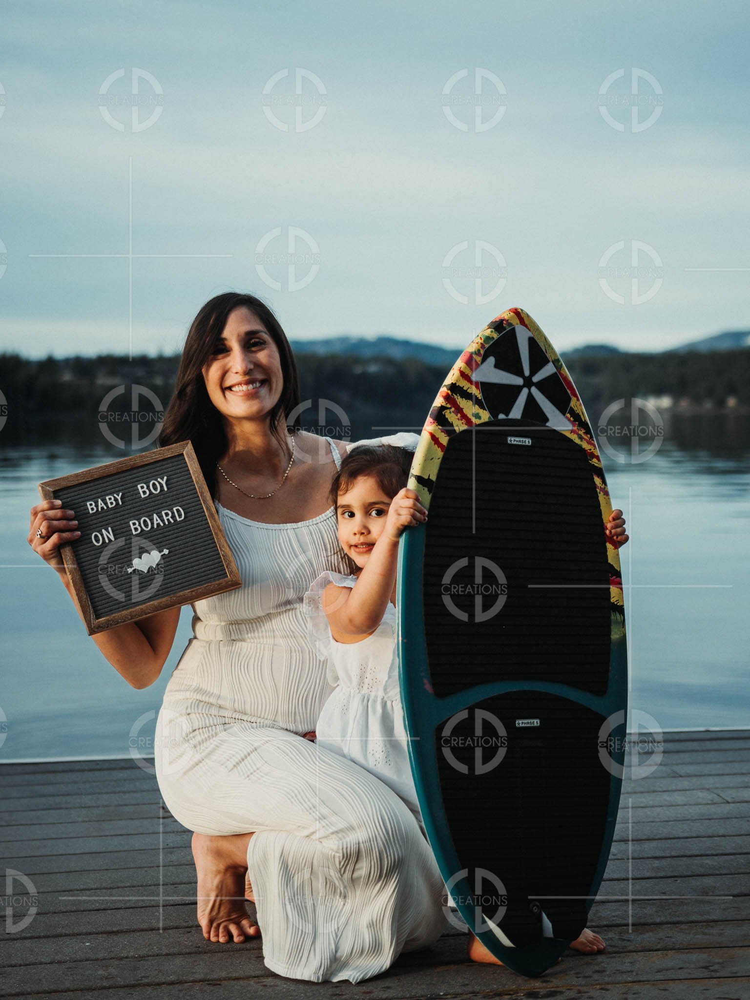 A woman and a young girl with a surfboard on a dock by the water, holding a sign that says 'Baby Boy on Board.'
