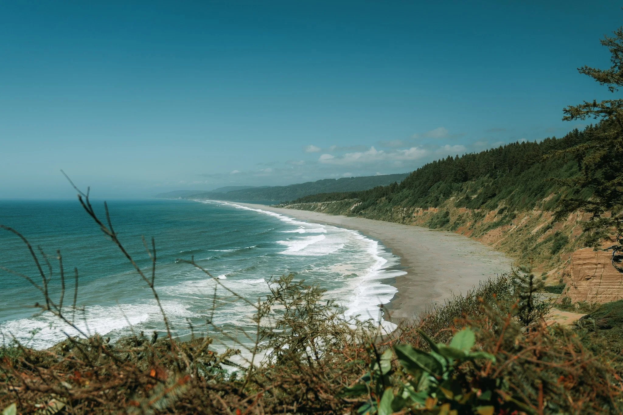 A scenic view of a coastline with sandy beach, blue ocean waves, green forested cliffs, and a partly cloudy sky.