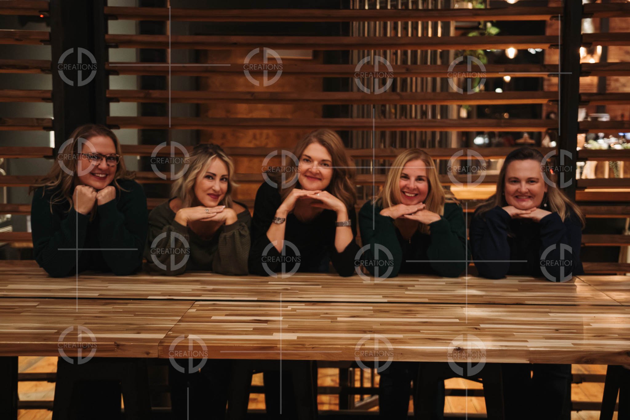 Five women sitting at a wooden table, smiling with their chins resting on their hands in a restaurant or cafe with wooden decor in the background.
