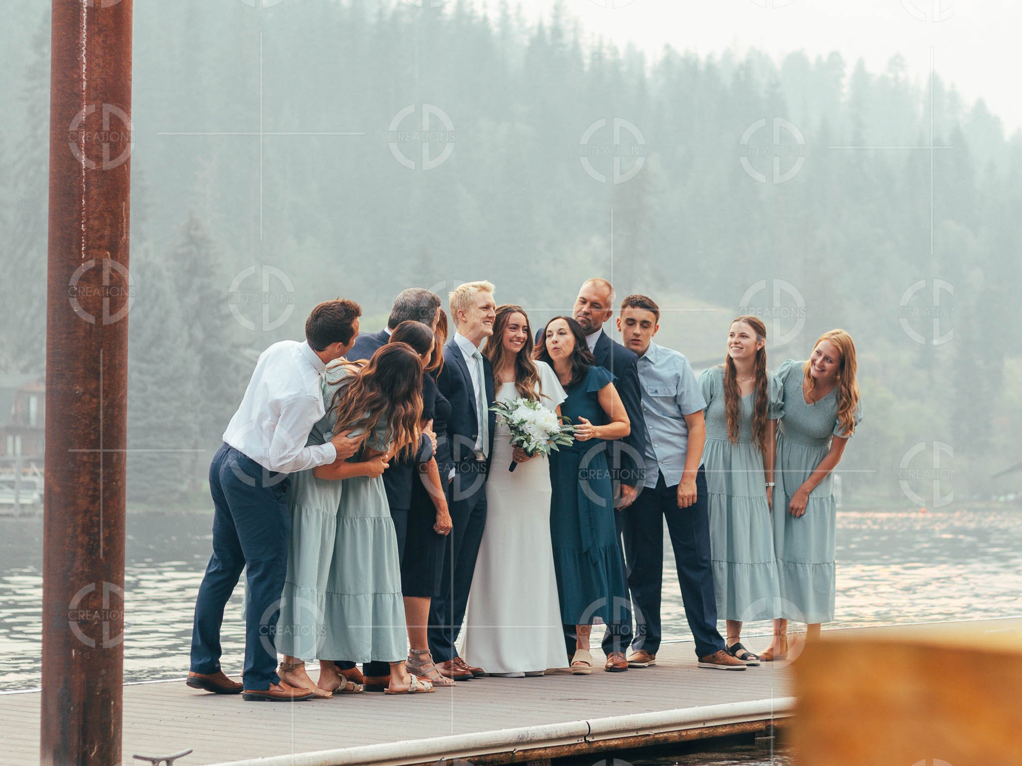 A group of people, including a bride holding a bouquet, gather on a dock by the water, smiling and posing for a photograph with a misty forest background.