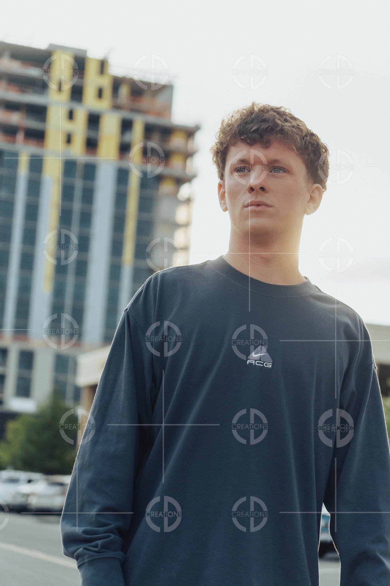Young man with curly hair and a long-sleeve black shirt standing outdoors with a modern building under construction and parked cars in the background.