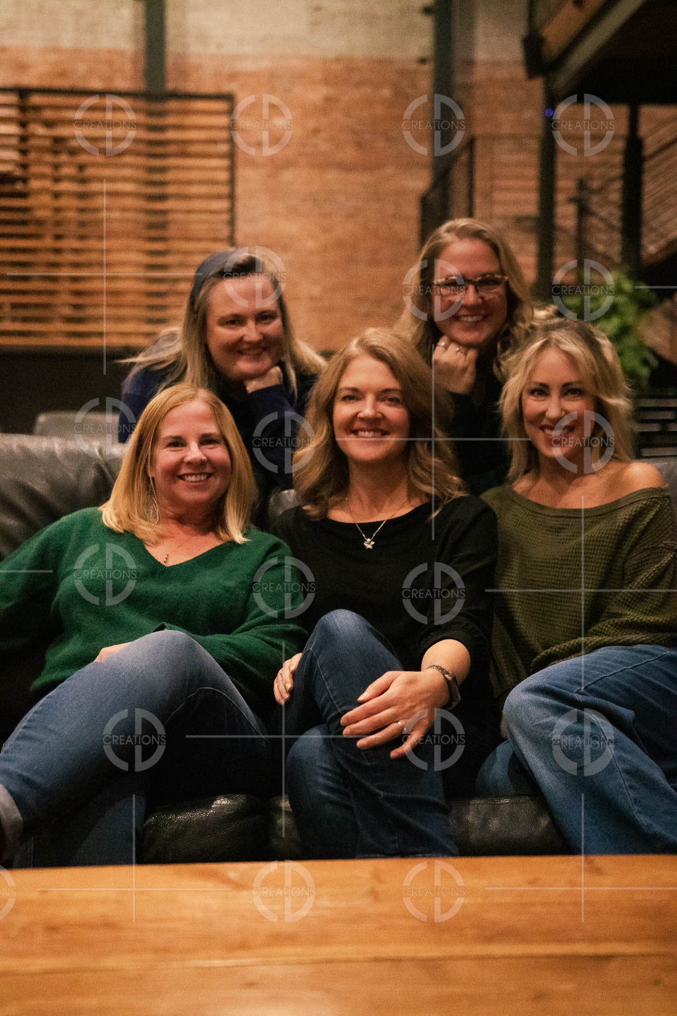 Group of five smiling women sitting on a leather couch in a cozy, indoor setting with brick walls and wooden accents.