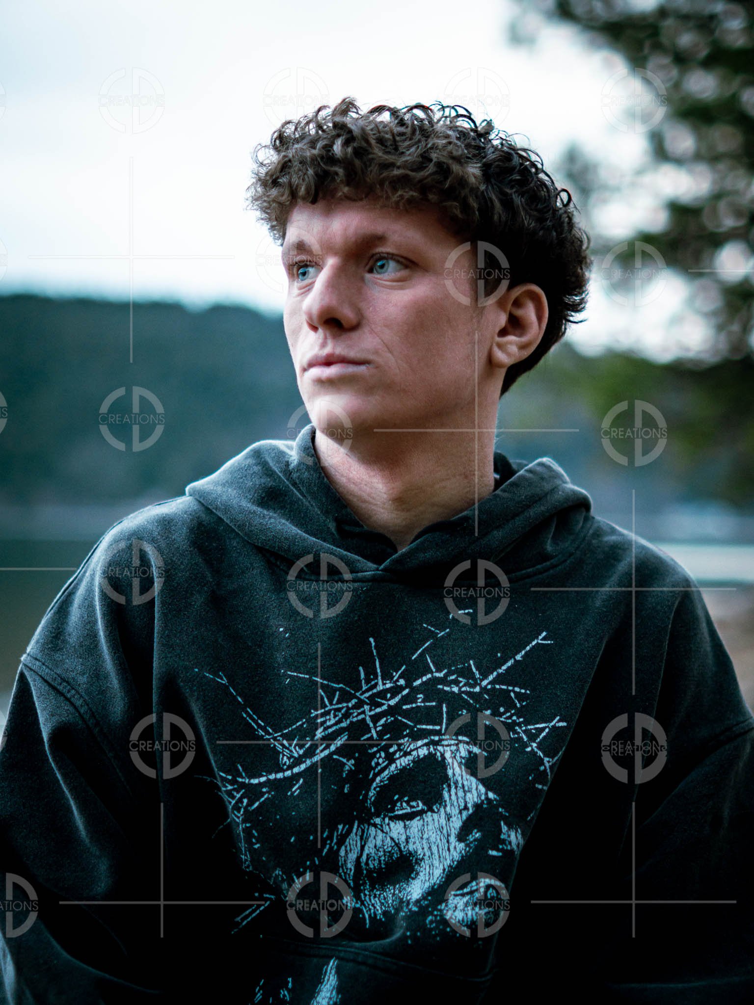 A young man with curly brown hair and blue eyes wearing a black hoodie with a graphic of a face and barbed wire on the front, standing outdoors with a blurred background of trees and sky.
