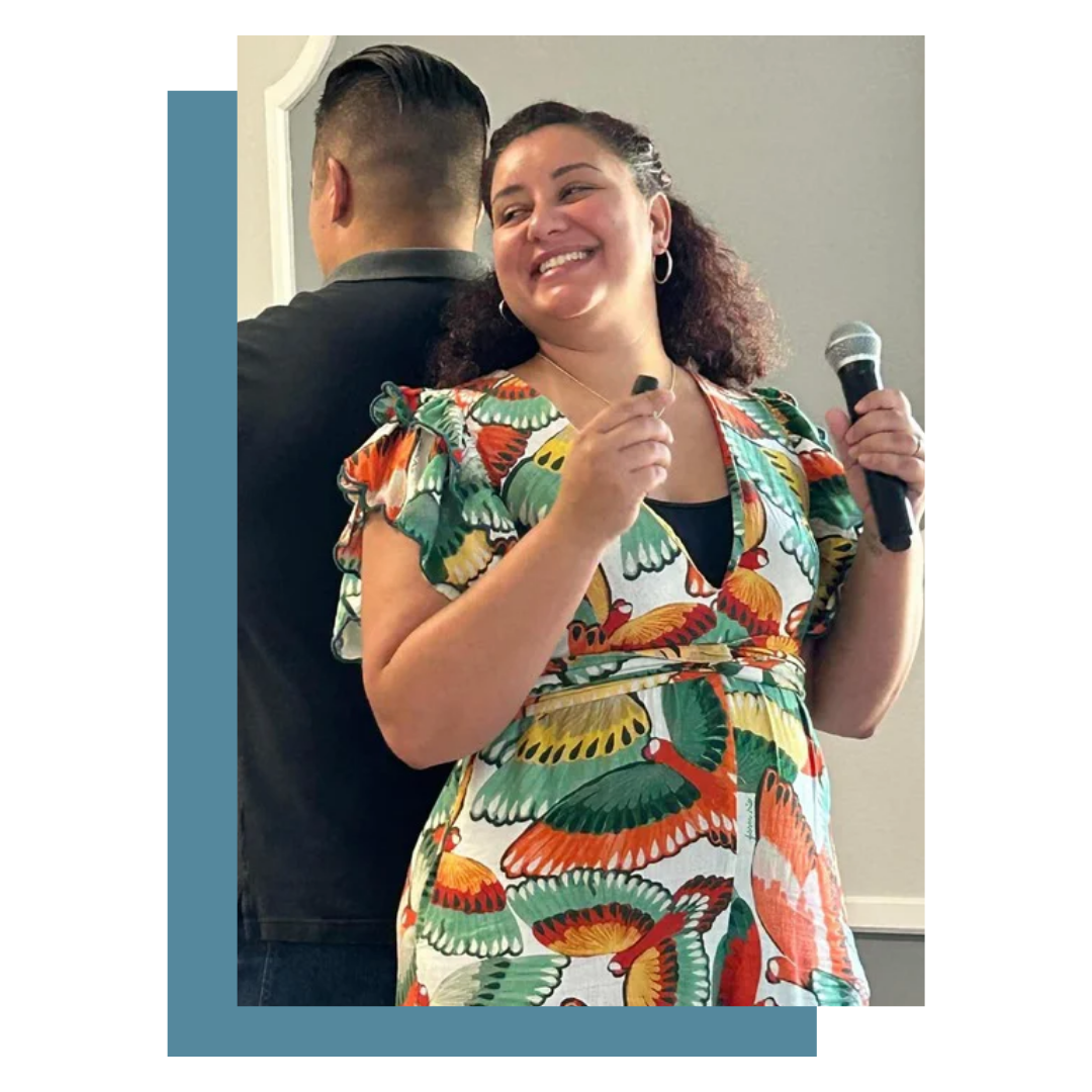 A woman smiling and holding a microphone, standing in front of a man with his back turned, wearing a colorful butterfly-patterned dress.