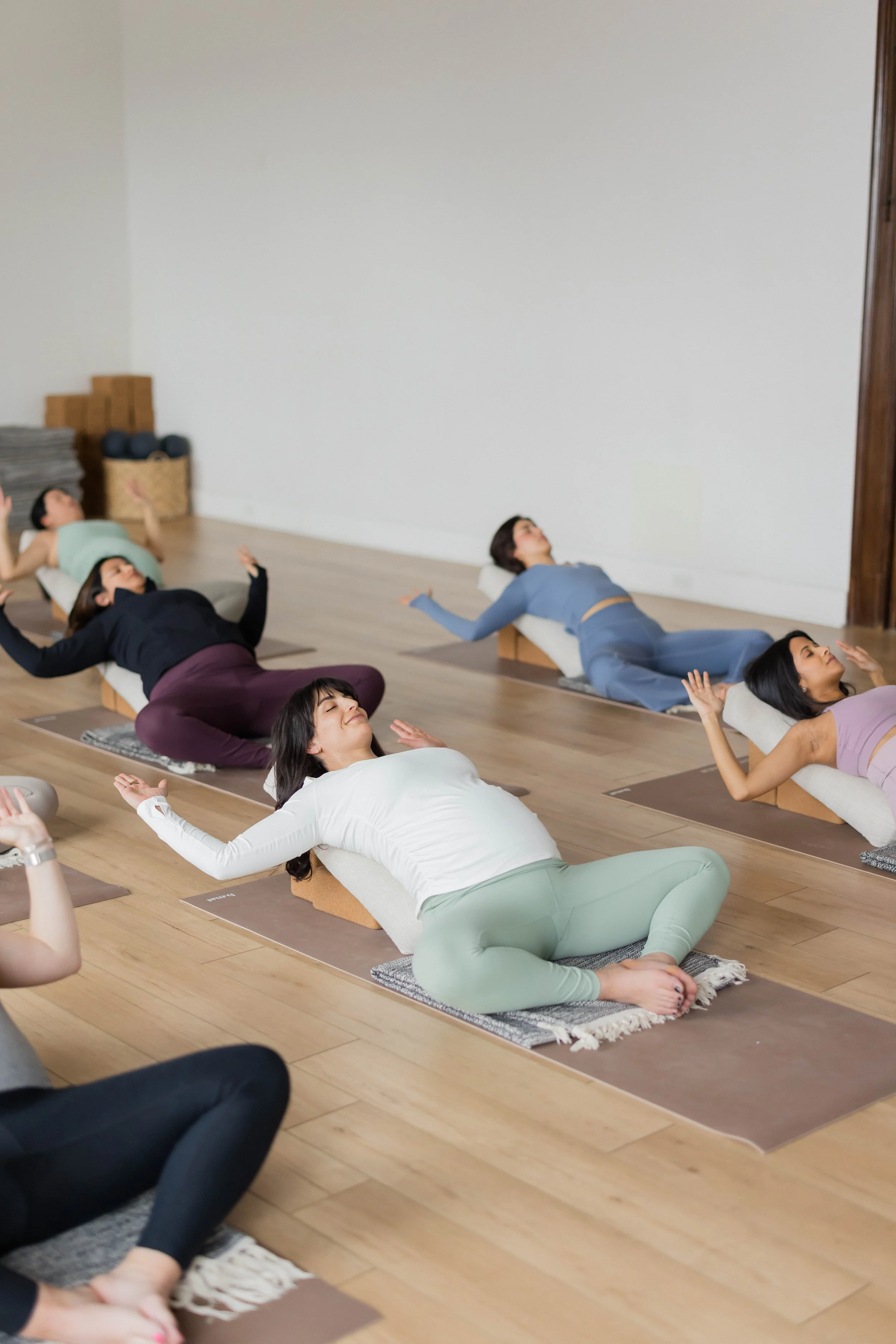 A group of women participating in a yoga class, lying on their yoga mats in a relaxed position with eyes closed, on a wooden floor in a studio with a plain white wall.