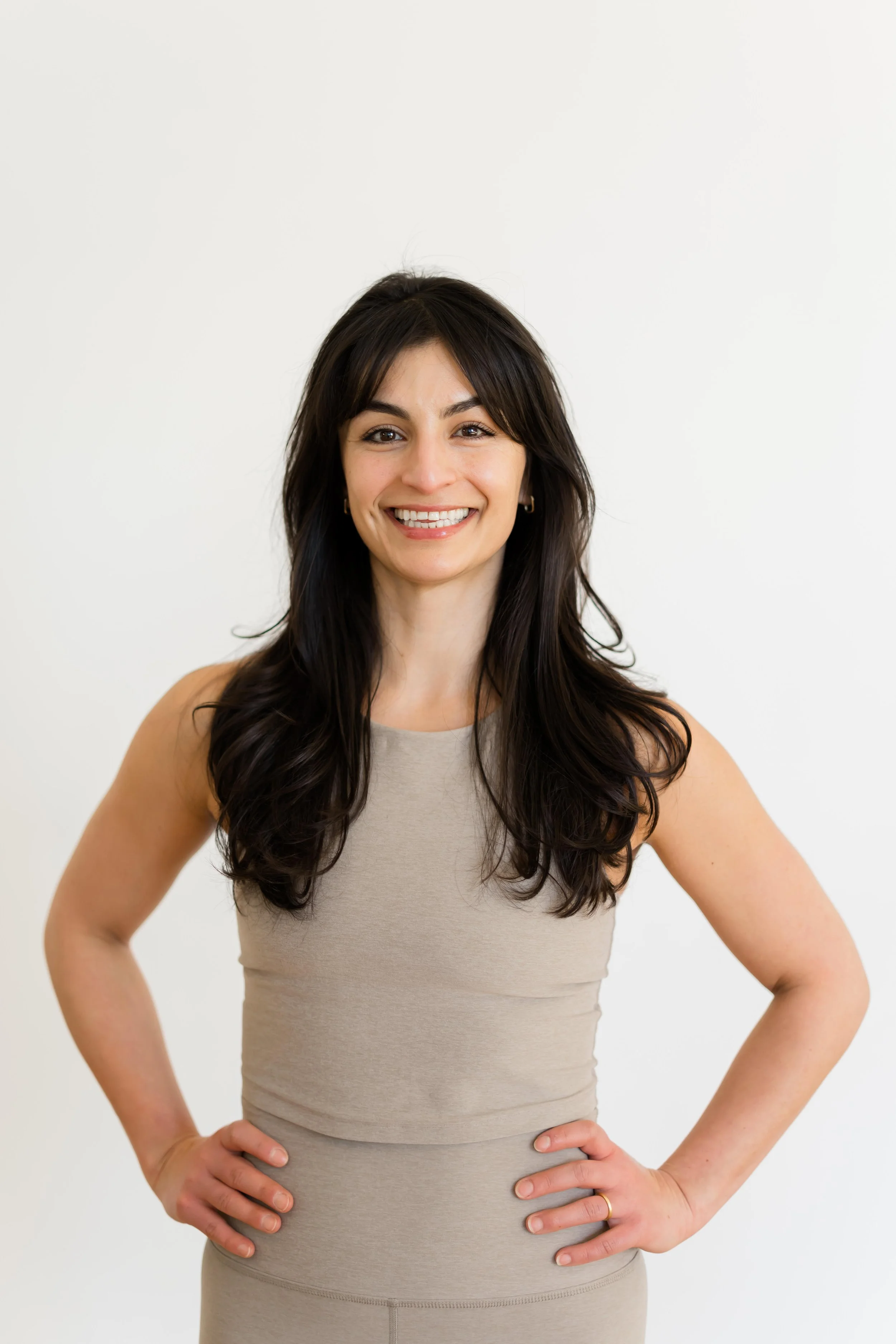 A woman with dark brown hair and a bright smile, wearing a sleeveless beige dress, standing against a plain white background with her hands on her hips.