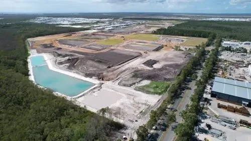 Aerial view of a large construction site with a body of water, ongoing earthwork, and surrounding commercial buildings and roads.