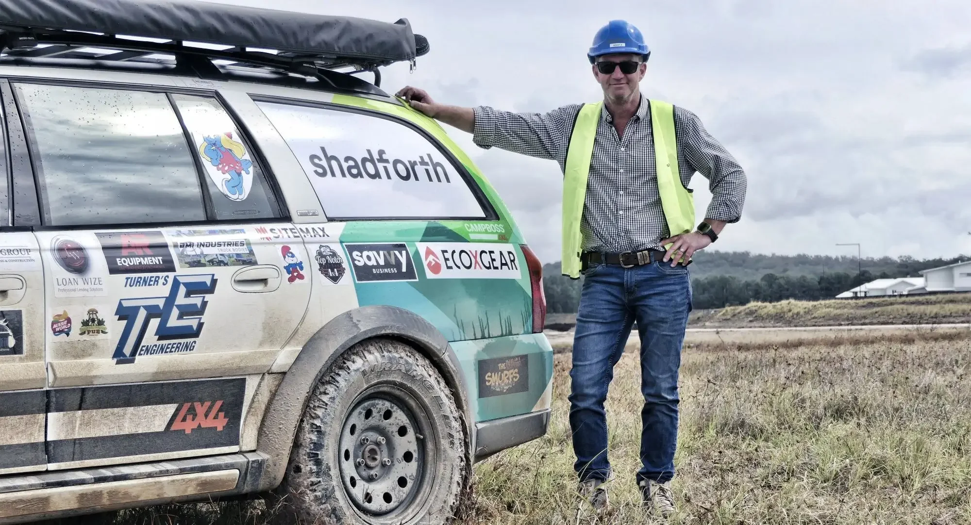 Man standing at the back of a charity car with the Shadforth logo. Man is wearing blue hard hat, yellow hi vis vest with blue jeans.