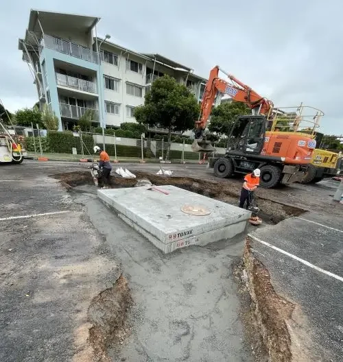Construction workers excavate around a large concrete utility box in a parking lot, with an orange excavator and a multi-story apartment building in the background.