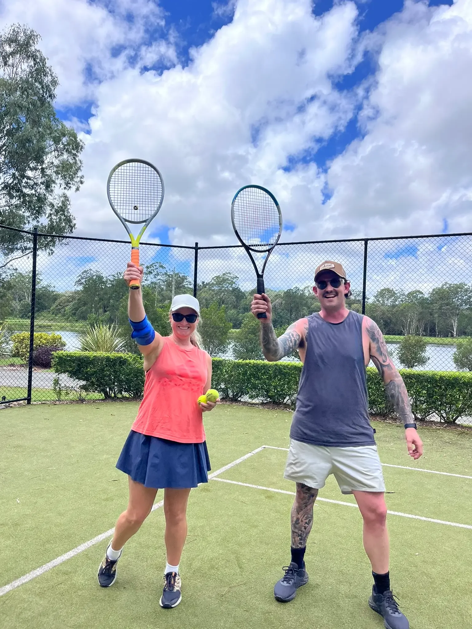 Two people standing on a tennis court holding up their tennis rackets smiling.