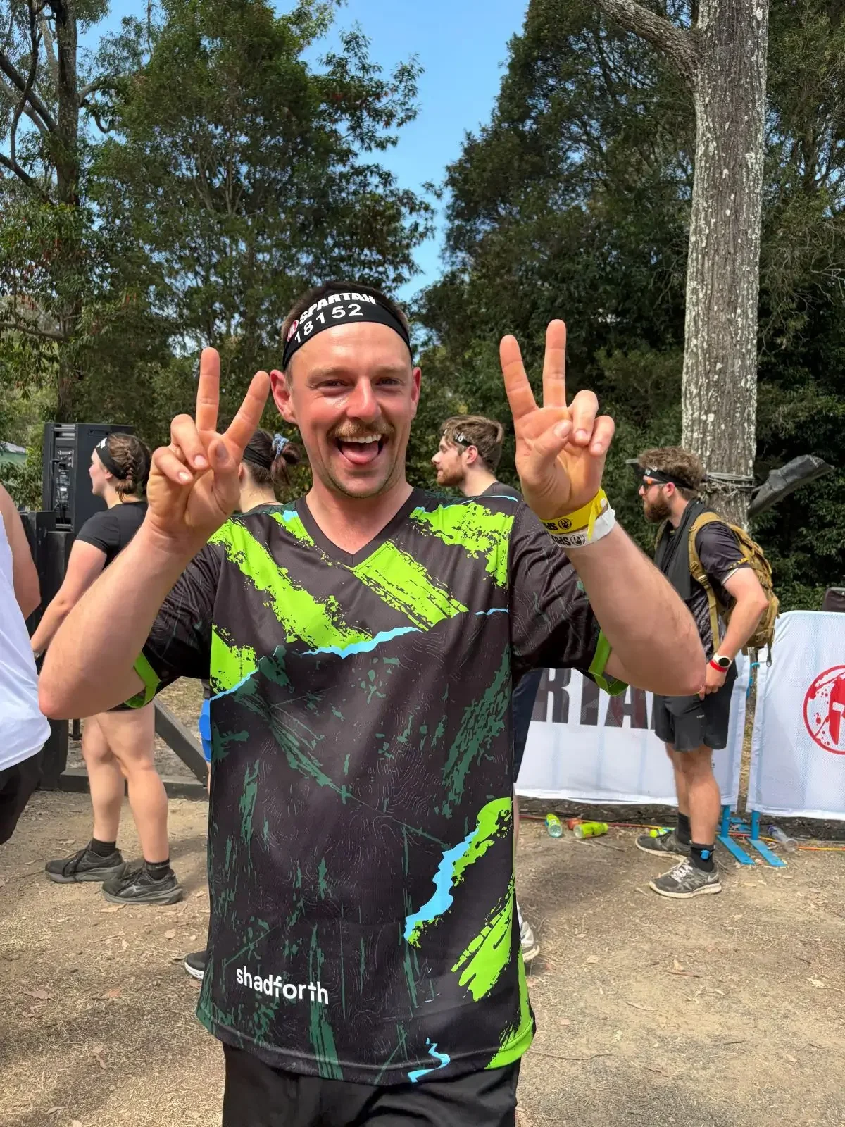 Man standing with his hands up smiling and wearing a green and black Shadforth running shirt. Trees and people in the background.