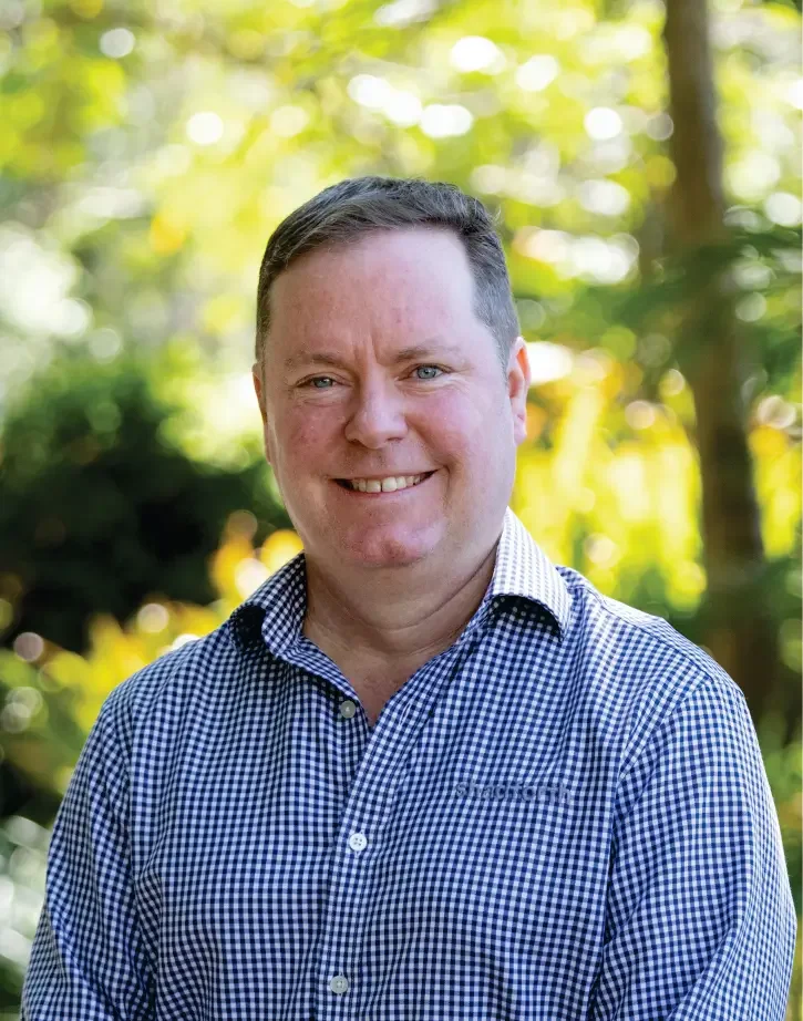 Chief Financial Officer Ben Baker smiling outdoors in a checkered blue and white shirt, with a blurred green and yellow background of trees and sunlight.
