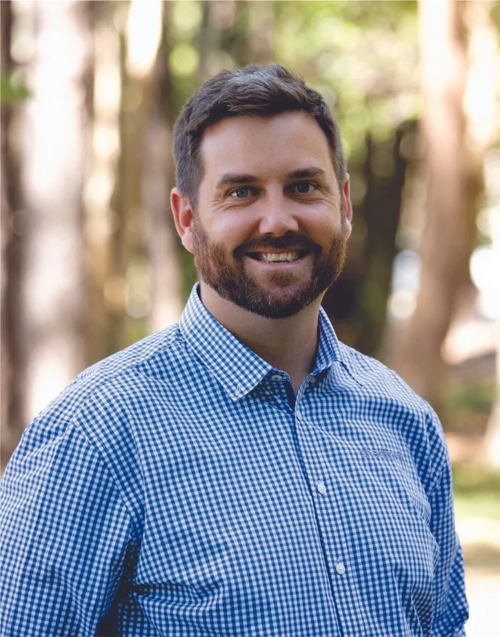 Executive Manager, Daniel Coonan wearing a blue and white checkered shirt, standing outdoors in a forested area with blurred trees in the background.