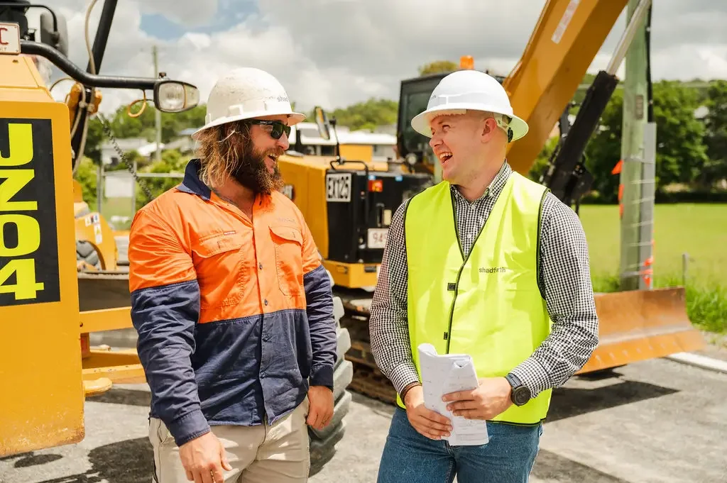 Two construction workers in white hard hats smile and talk on site, with construction equipment and trees in the background.