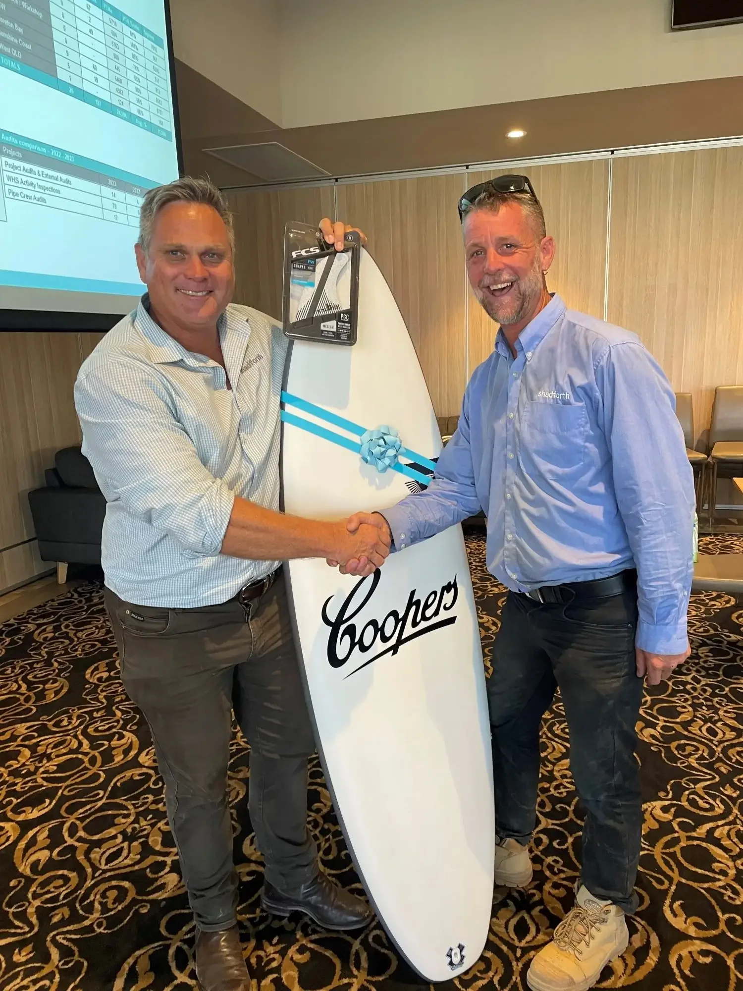 Two people standing in a room shaking hands while holding a surfboard with a blue bow on it.