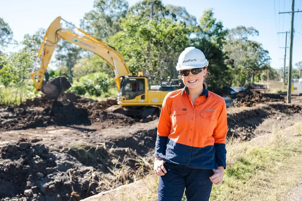 A smiling woman wearing hi vis construction standing outdoors near a construction site, with an excavator digging in the background.