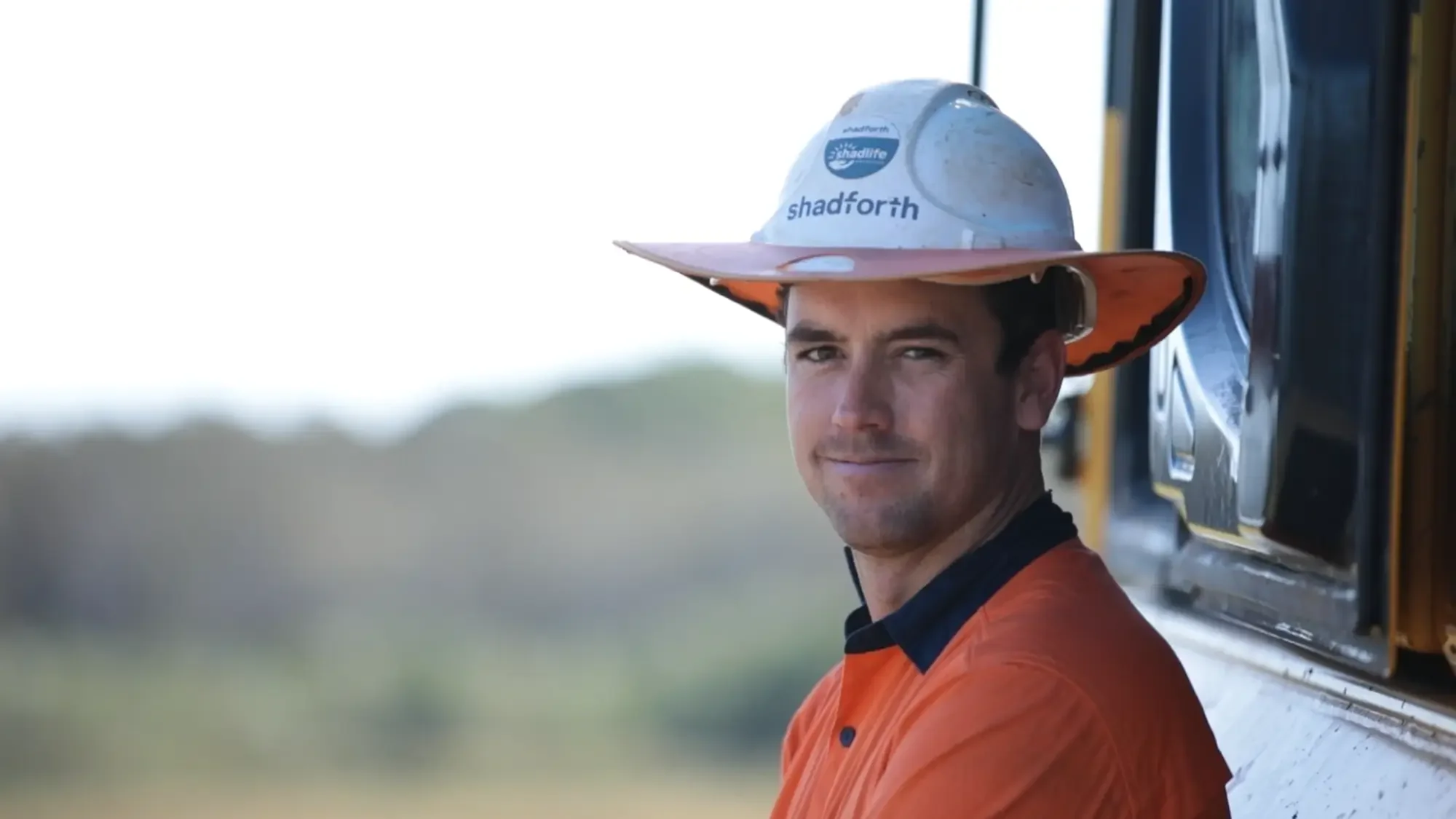 A construction worker in an orange hi vis shirt and a hard hat leaning against machinery.