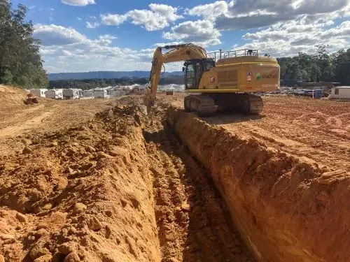 Excavator working on a construction site digging a trench in the dirt under a partly cloudy sky.