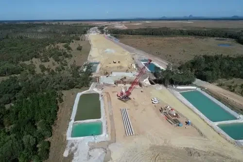 Aerial view of a construction site with a crane, two rectangular pools, and ongoing road or infrastructure development in a rural area with trees and open land.