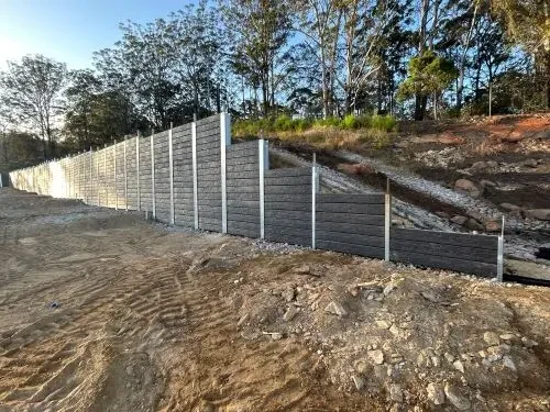 A constructed retaining wall made of dark panels with white support posts on a sloped terrain, with trees and a clear sky in the background.