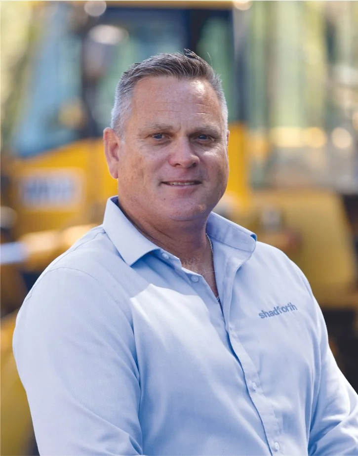 Executive General Manager, Stuart Dodunski in light blue shirt with Shadforth logo, standing outdoors with yellow construction vehicle in background.