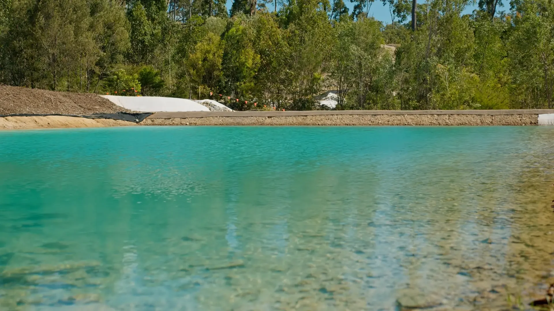 Calm turquoise body of water with a shoreline that has dirt and white plastic sheeting in front of a dense green forest.