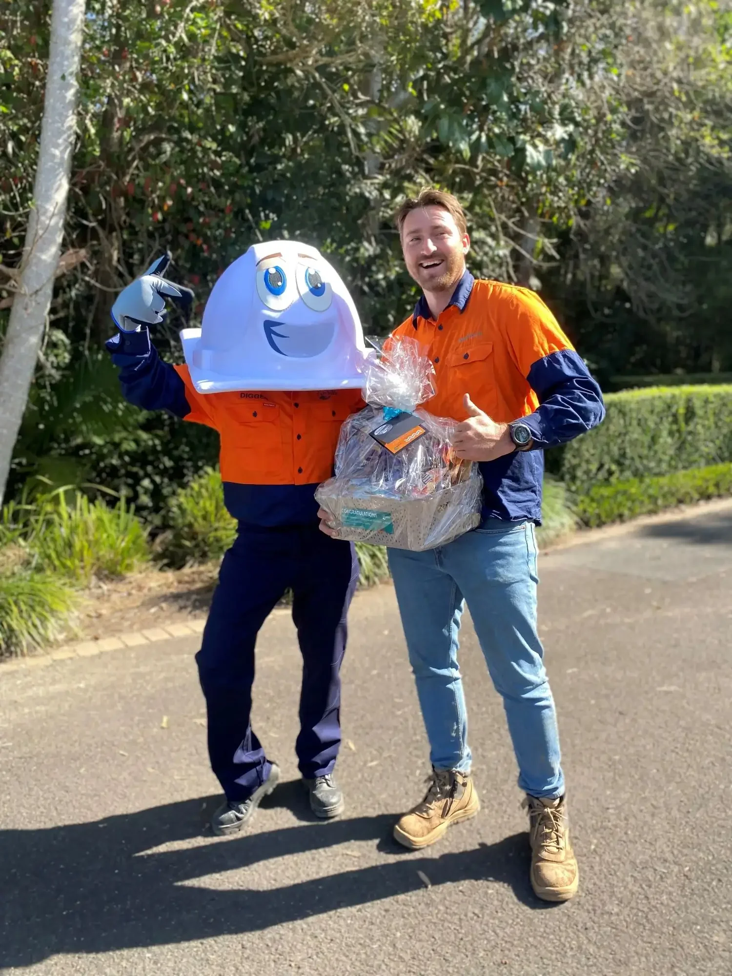 A man in a construction uniform smiling with his thumb up holding a hamper. He is standing next to a mascot.