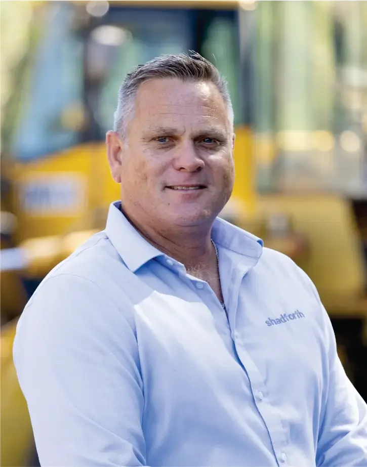 Executive General Manager, Stuart Dodunski in light blue shirt with Shadforth logo, standing outdoors with yellow construction vehicle in background.