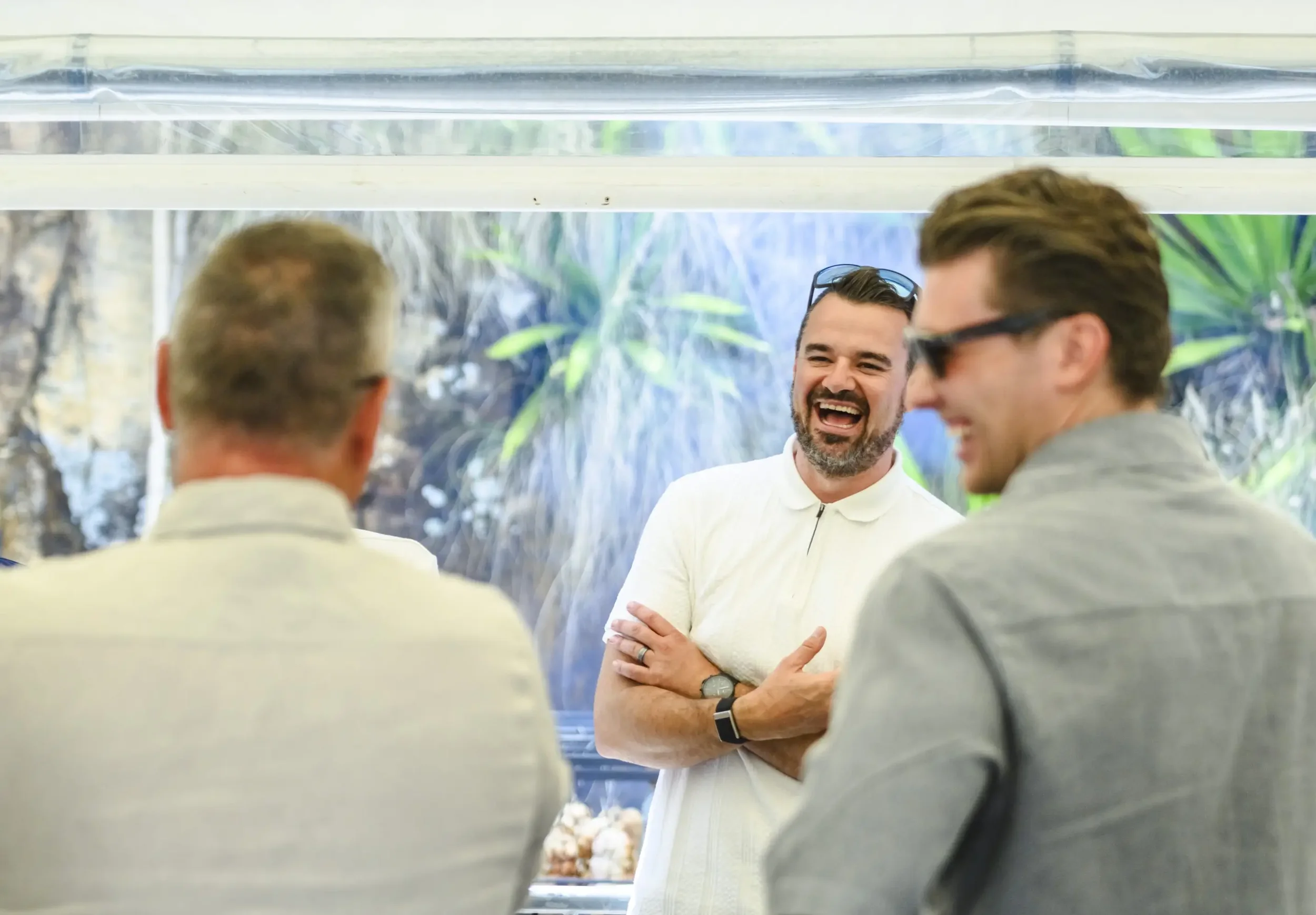Three men engaged in conversation indoors with a large decorative waterfall wall behind them, smiling and laughing.