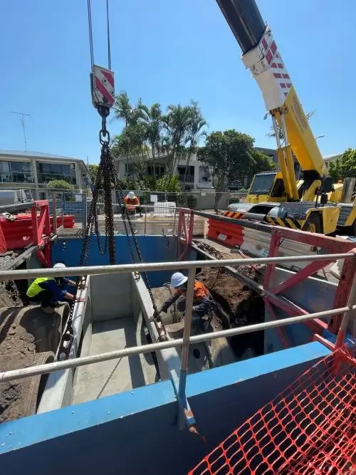 Construction workers working inside a large concrete structure at a construction site. A yellow crane is lifting a section into place, and there is protective barriers around the site.