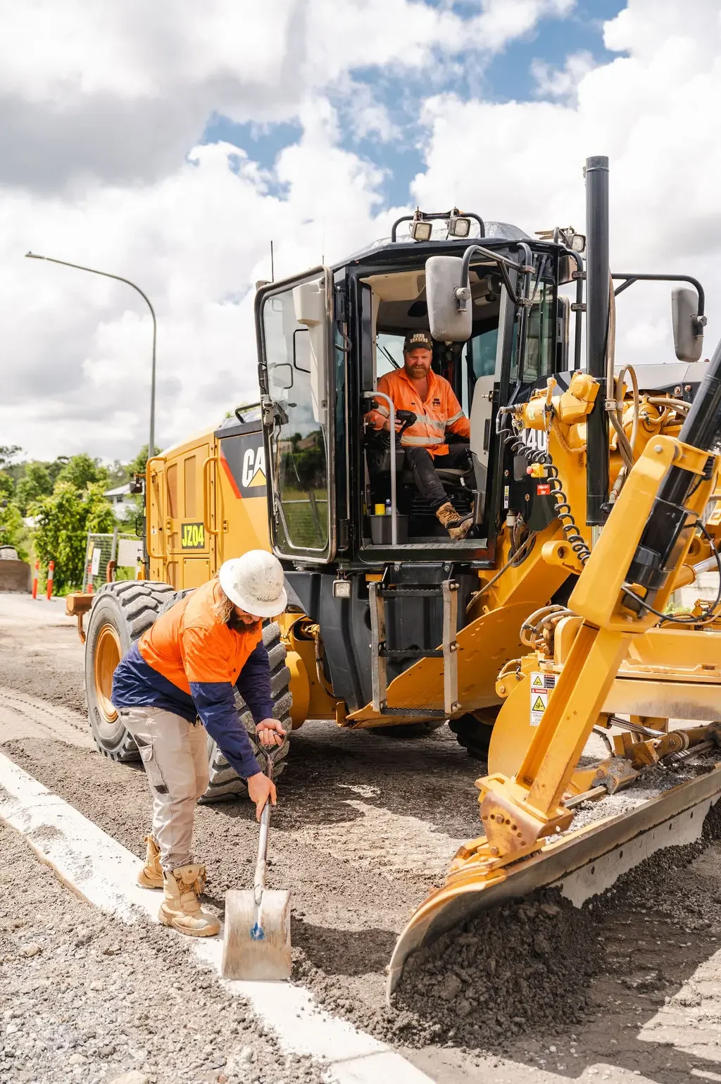 Construction workers working on a road with a yellow scraper. One worker is in the scraper, and another is shoveling gravel along the edge of the road.