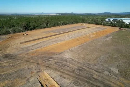 A large cleared construction site with dirt and gravel, surrounded by trees and industrial buildings in the distance, under a clear blue sky.