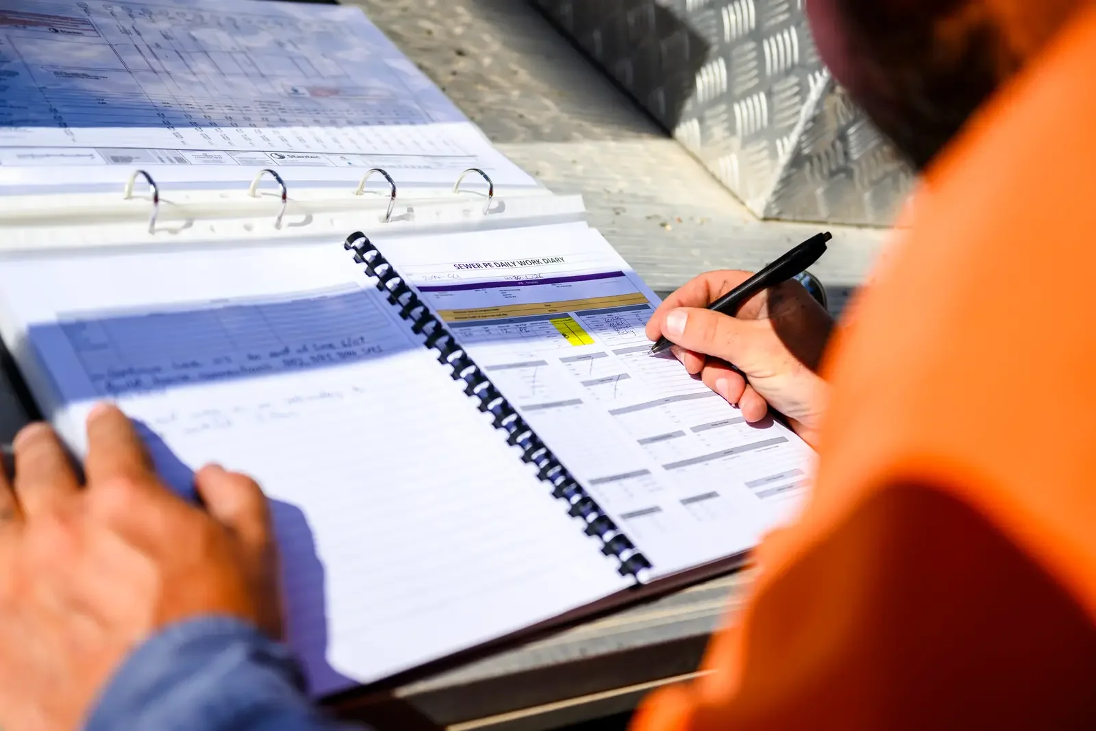 Person filling out paperwork at an outdoor table with various forms and documents, wearing an orange shirt.