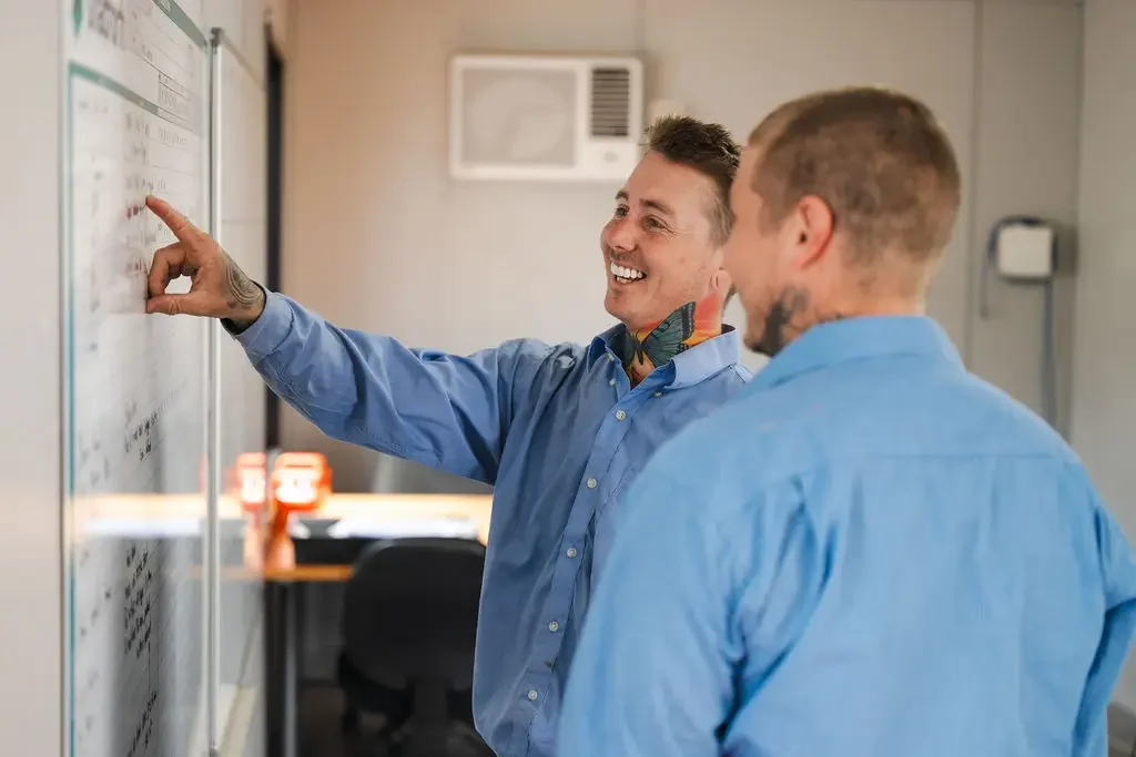Two men wearing blue shirts are smiling and discussing something on a whiteboard in an office setting.
