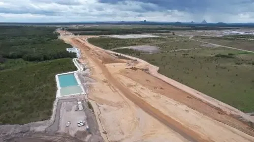 Partially built Bells Creek Arterial, some parked cars, and a water retention pond, surrounded by green fields and forests, under a cloudy sky.