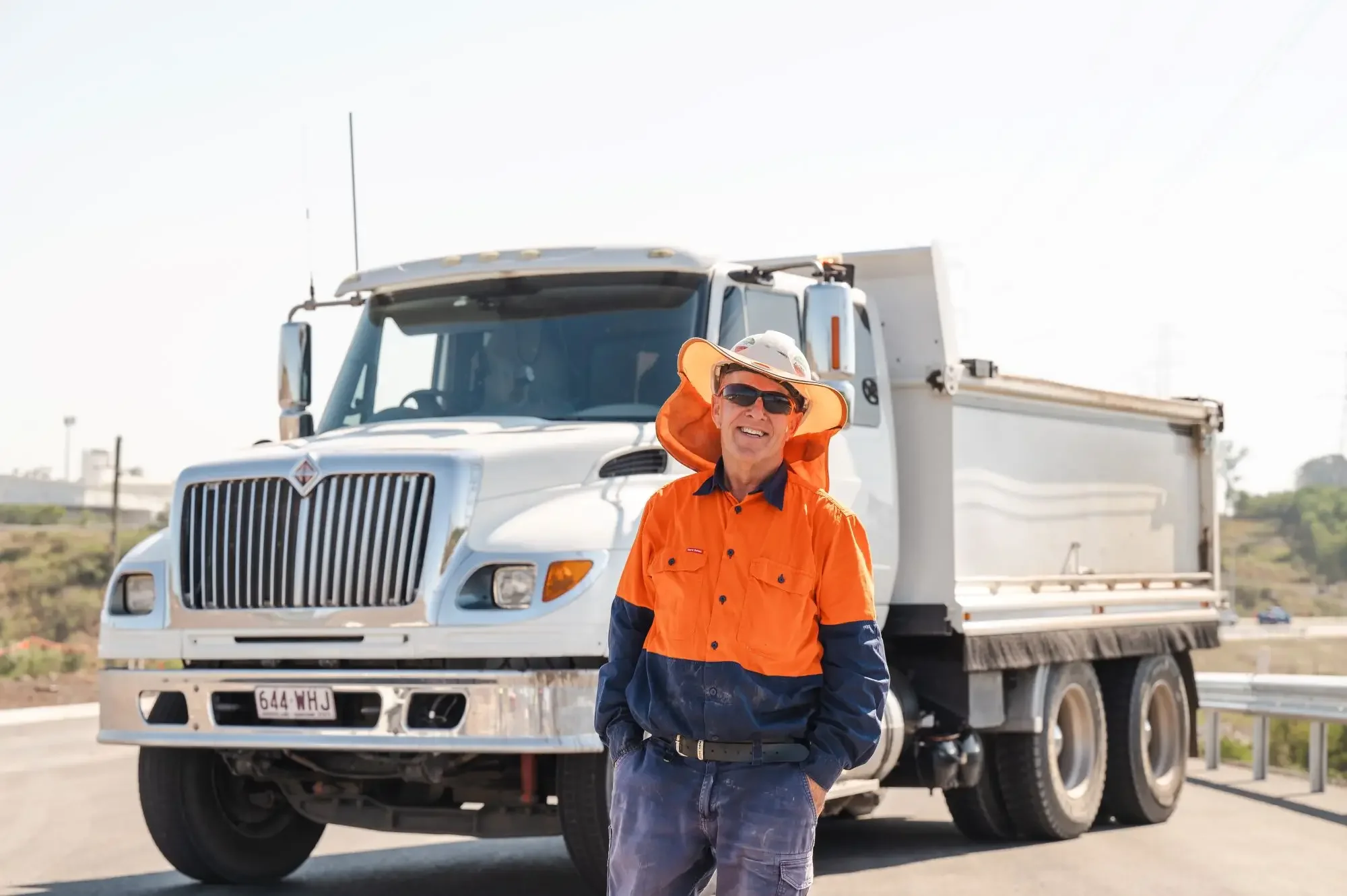 A man in orange and navy hi vis uniform and hard hat standing in front of a large white semi-truck on a highway during daytime.