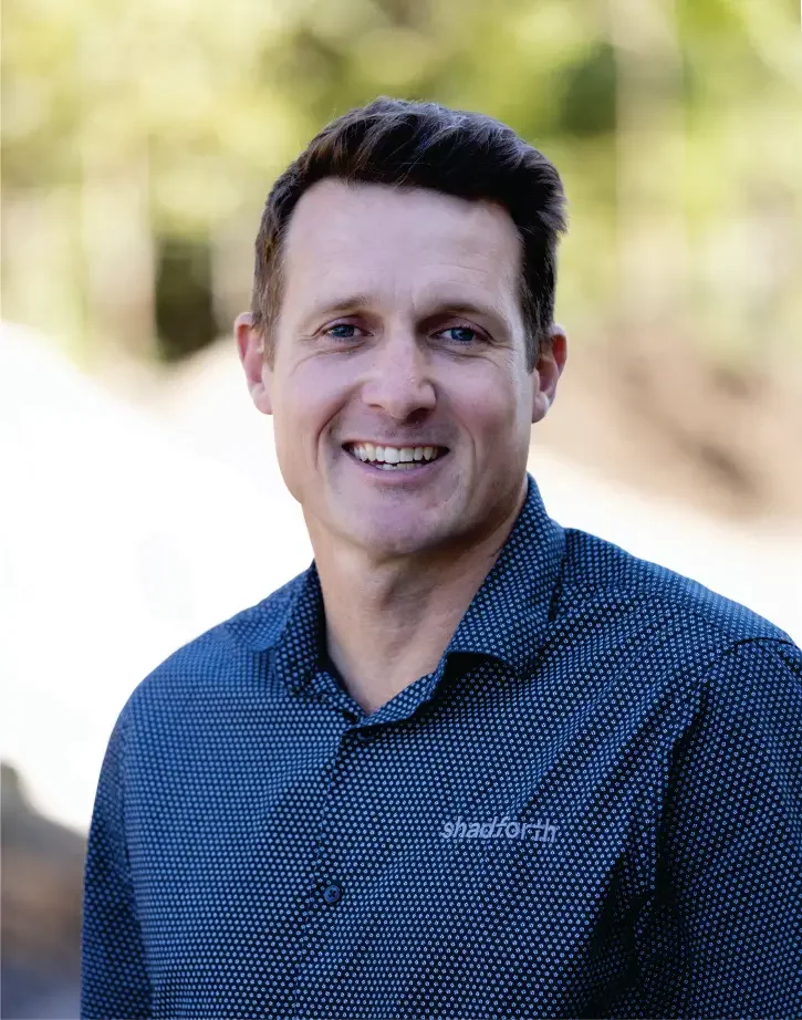 Executive General, Manager Michael Bishop wearing a blue patterned shirt outdoors with blurred green foliage in the background.