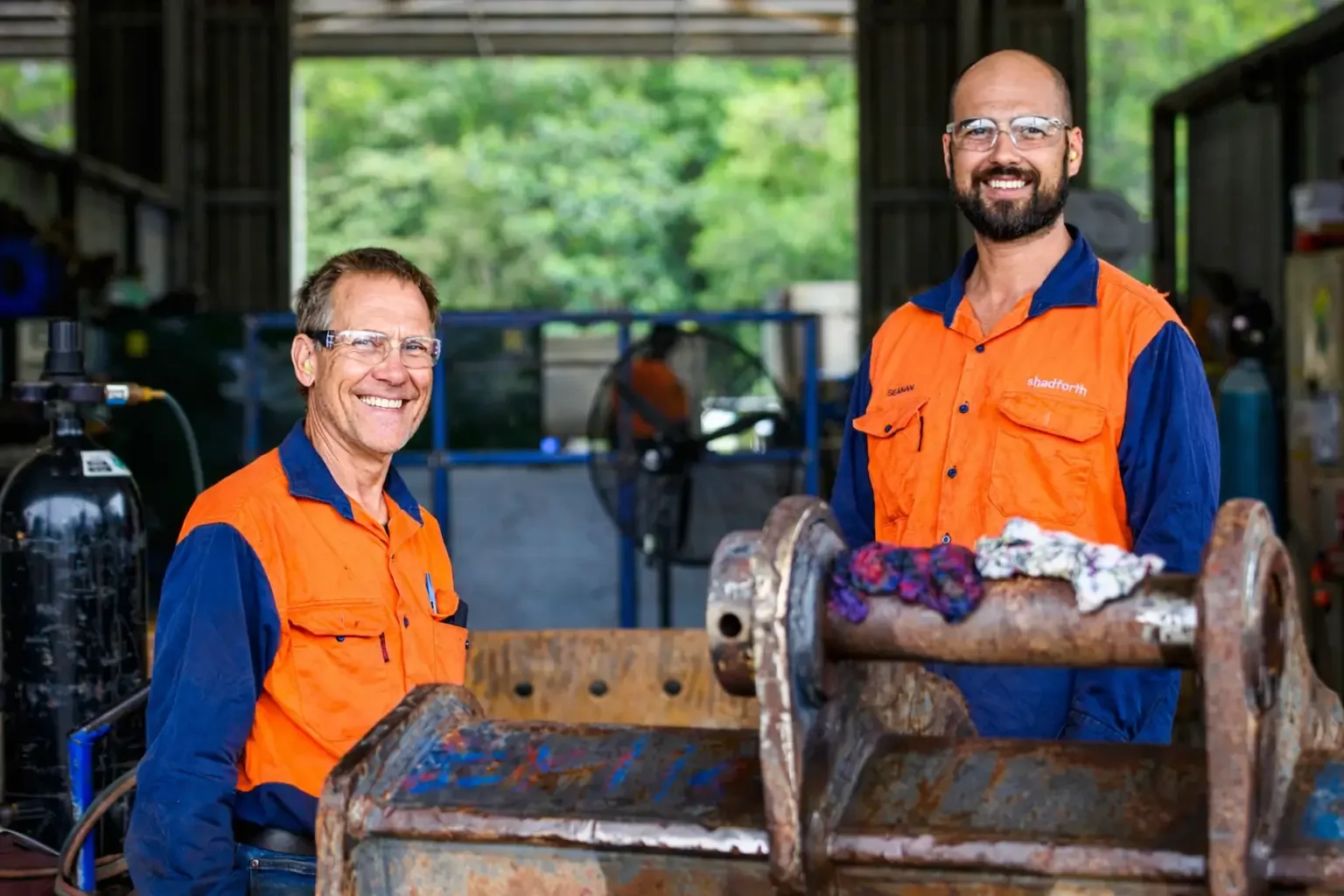Two men in orange and blue work shirts smiling inside a workshop with equipment and tools, working on a mechanical component.