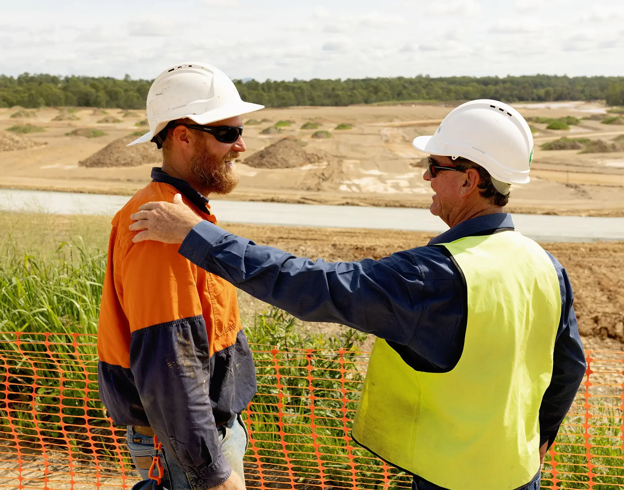 Two construction workers, shaking hands on a construction site near a body of water, with a landscape of dirt mounds and green trees in the background.