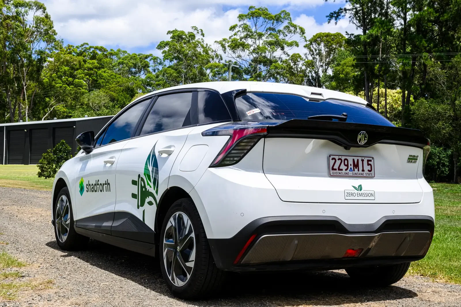 White hybrid car with the Shadforth logo and sustainable sticker. Driving on a gravel road with trees in the background.