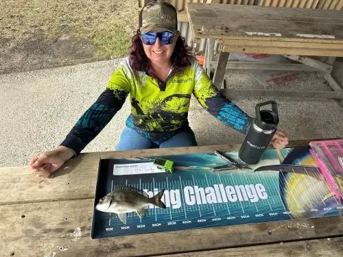 A lady sitting at a table wearing a Shadforth fishing shirt and hat. She has a fish on her brag mat.