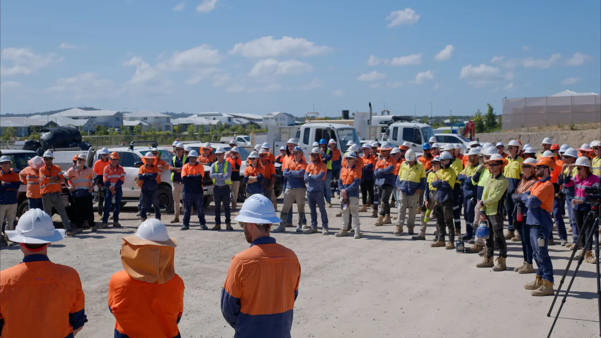 Group of construction workers standing outdoors on a construction site, wearing safety gear, with vehicles and construction equipment in background.