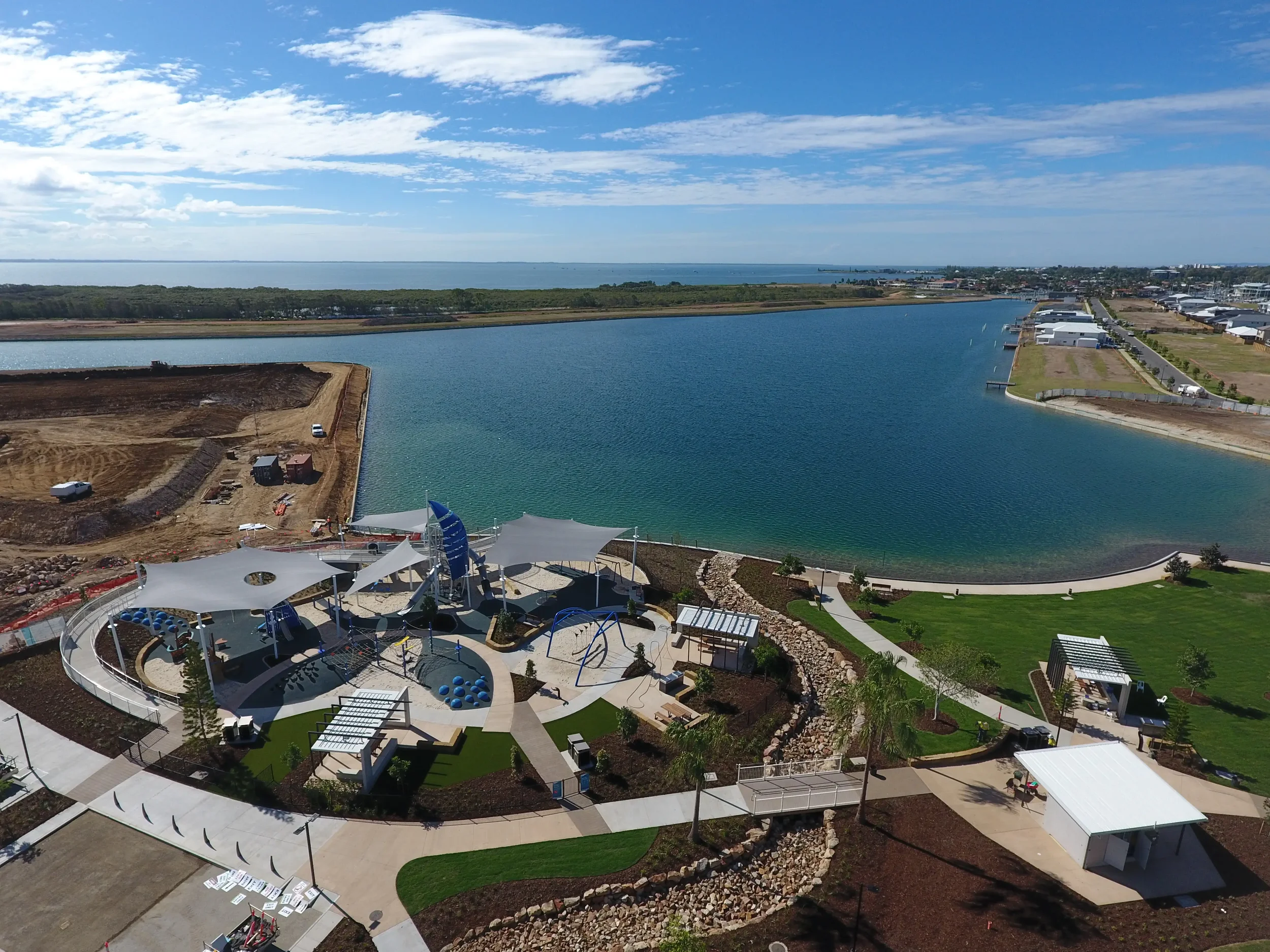 Aerial view of a waterfront park at Newport under construction with a large body of water, a marina, and a distant city skyline under a partly cloudy sky.