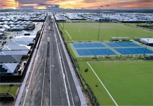 Image of houses on the left, an unfinished road and blue netball court and green fields on the right.